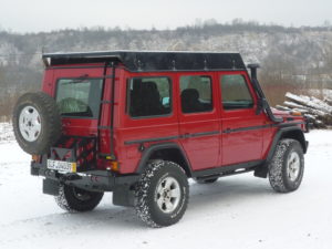Red G-Wagen with rear swinging tire mount and fuel tank, side rails, snorkel, and roof rack.
